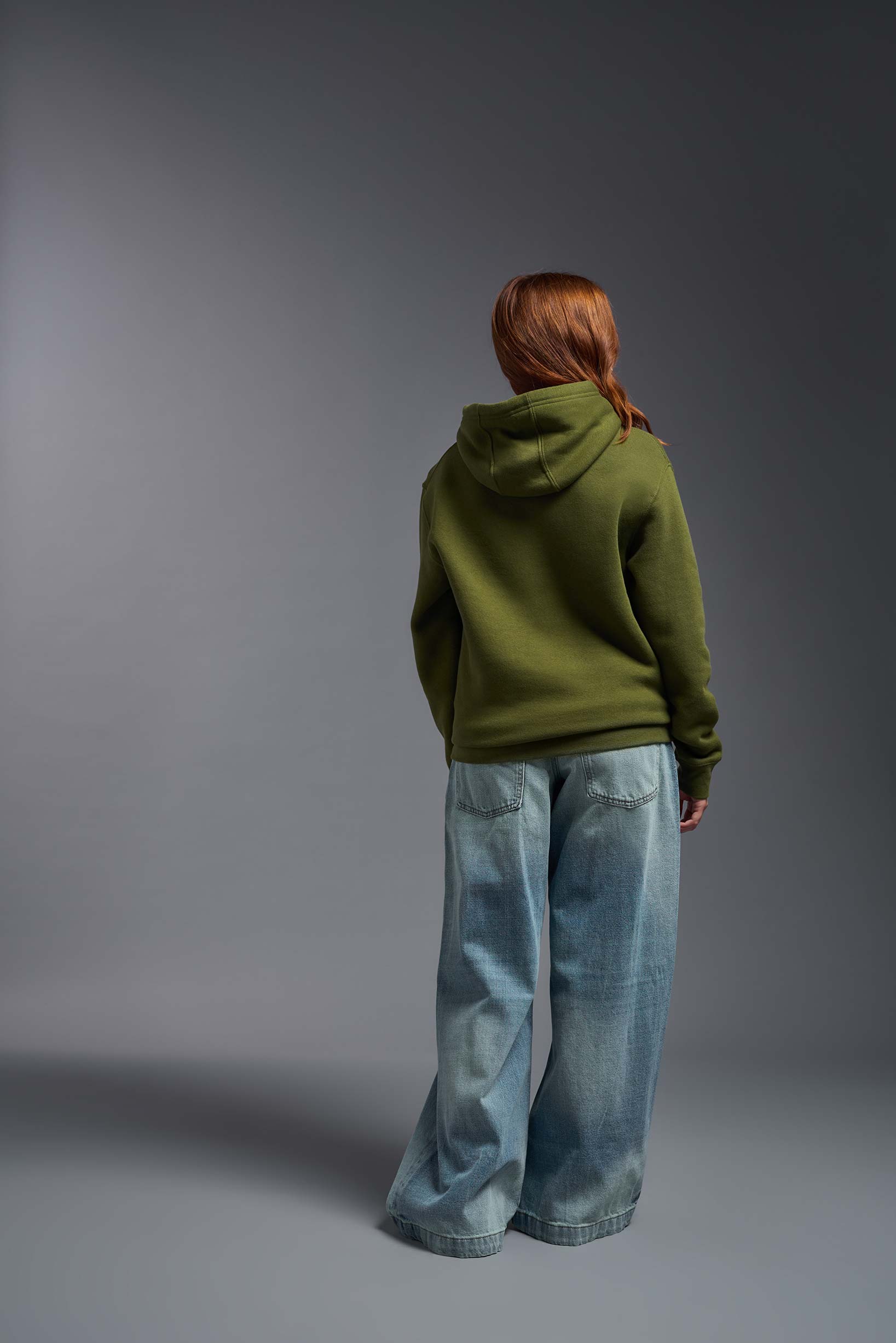 A female model wearing the unisex Americana 3-End Fleece Pullover Hoodie in the color military green, made from 75% combed ring-spun USA-grown cotton and 25% polyester, showing the relaxed fit from a back view. This premium fleece hoodie is Made in the USA. #military-green
