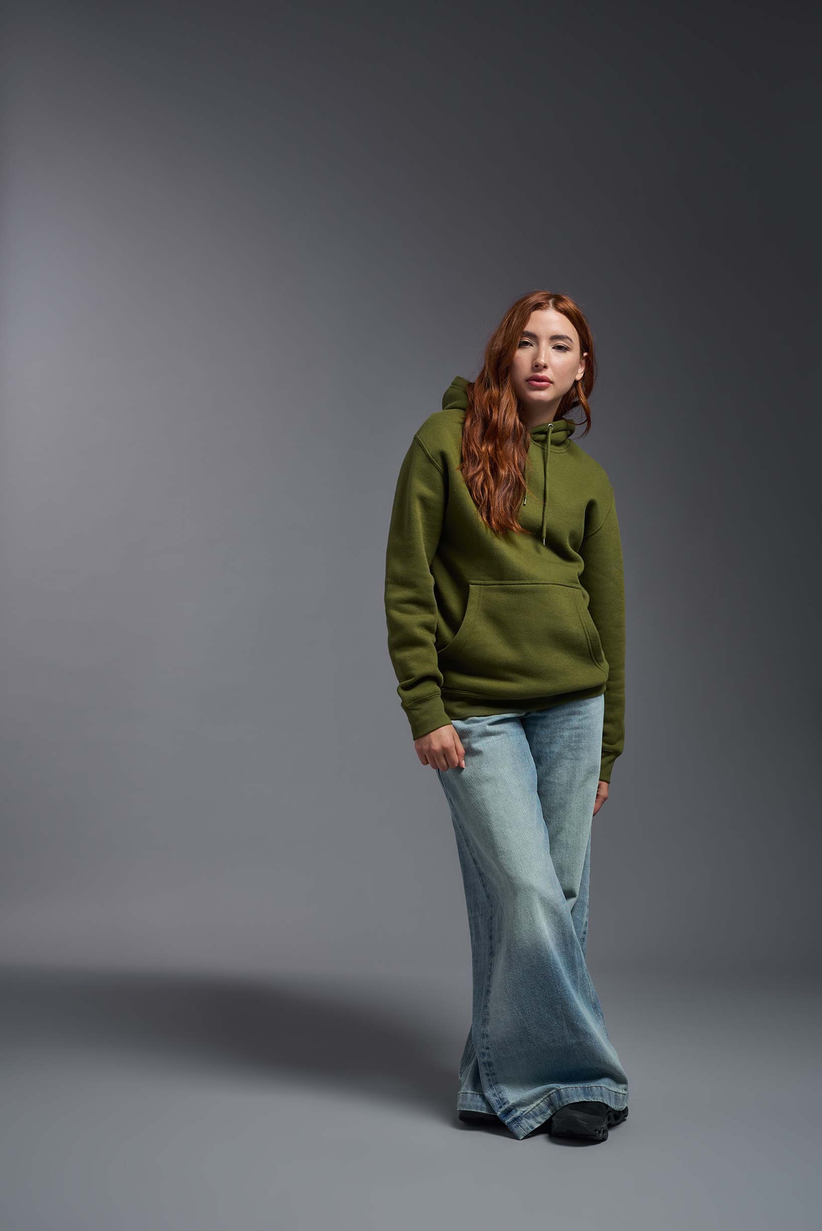 A female model wearing the unisex Americana 3-End Fleece Pullover Hoodie in the color military green, made from 75% combed ring-spun USA-grown cotton and 25% polyester, showing the relaxed fit from a front view. This premium fleece hoodie is Made in the USA. #military-green