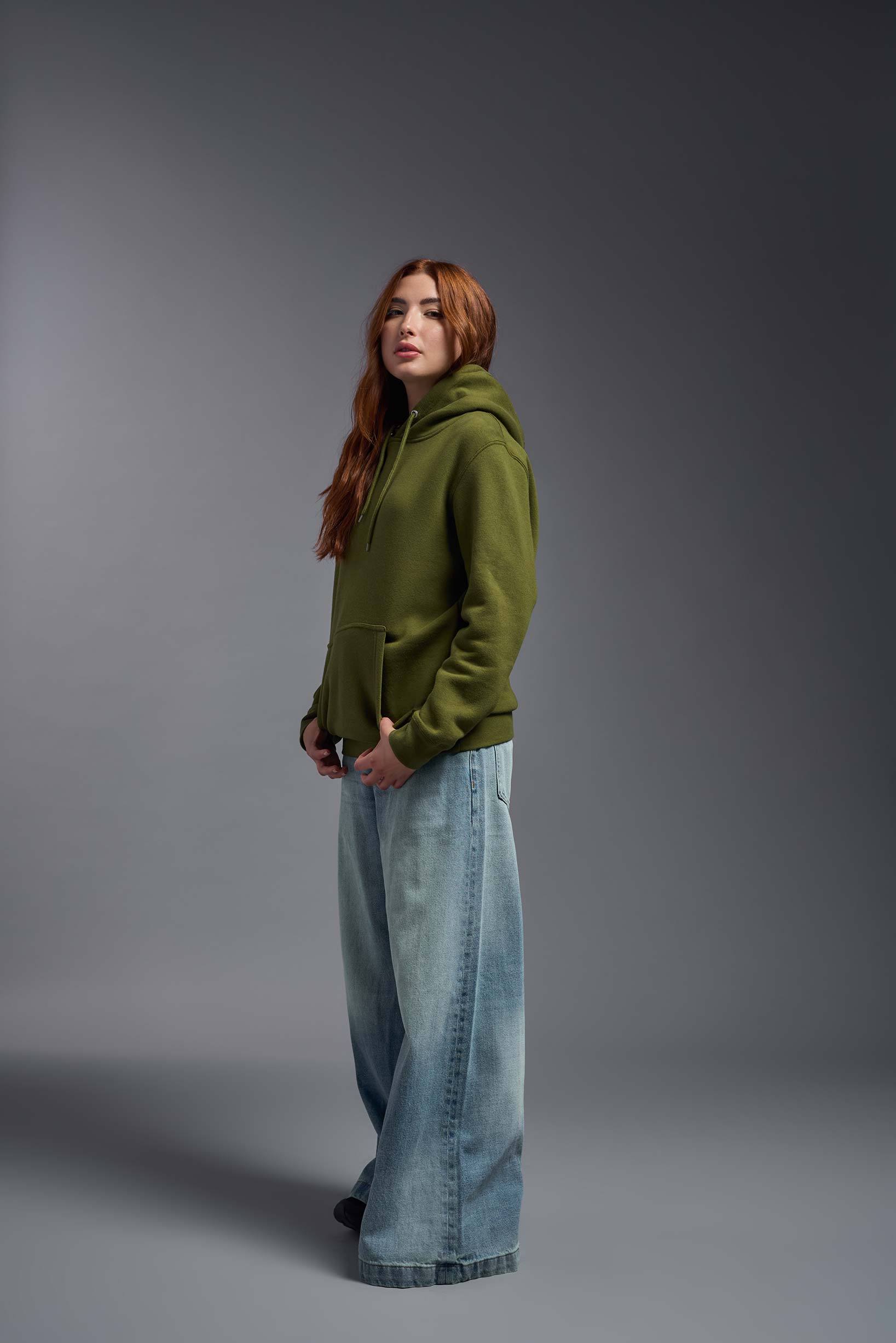 A female model wearing the unisex Americana 3-End Fleece Pullover Hoodie in the color military green, made from 75% combed ring-spun USA-grown cotton and 25% polyester, showing the relaxed fit from a side view. This premium fleece hoodie is Made in the USA. #military-green