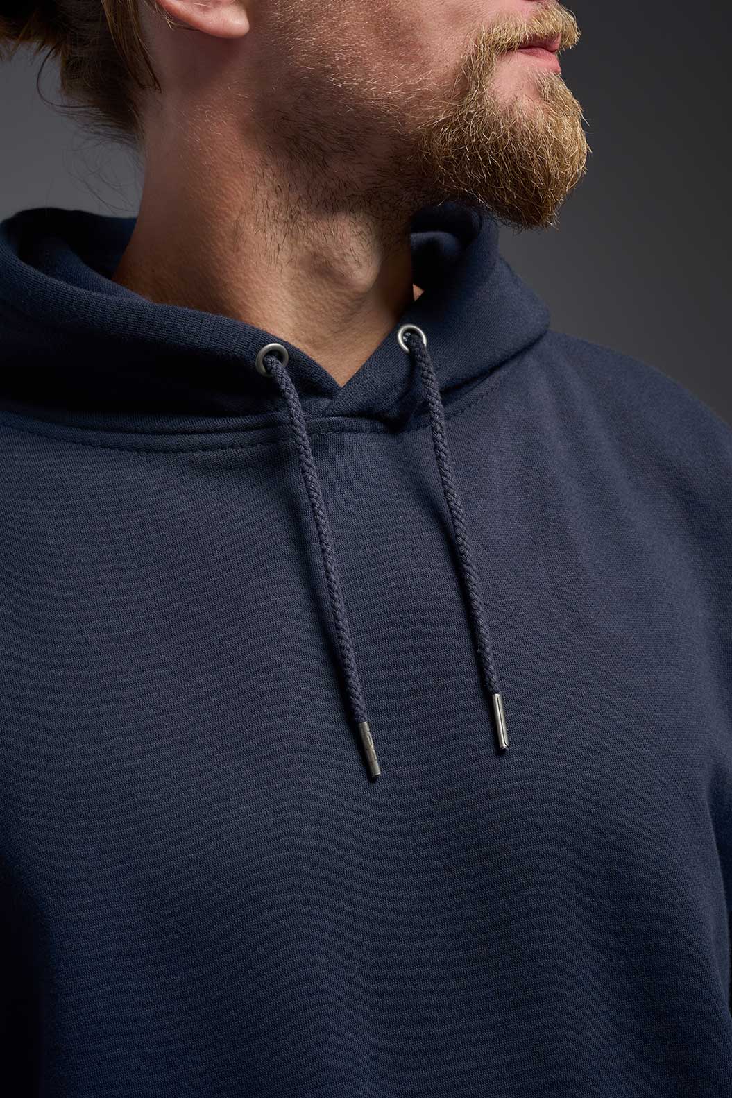 A male model wearing the unisex Americana 3-End Fleece Pullover Hoodie in the color navy, made from 75% combed ring-spun USA-grown cotton and 25% polyester, showing the relaxed fit from a close-up collar view. This premium fleece hoodie is Made in the USA. #navy