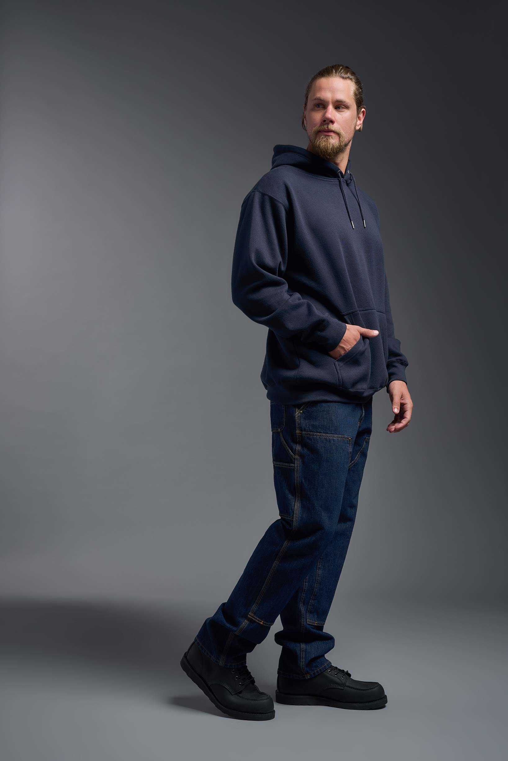 A male model wearing the unisex Americana 3-End Fleece Pullover Hoodie in the color navy, made from 75% combed ring-spun USA-grown cotton and 25% polyester, showing the relaxed fit from a side view. This premium fleece hoodie is Made in the USA. #navy