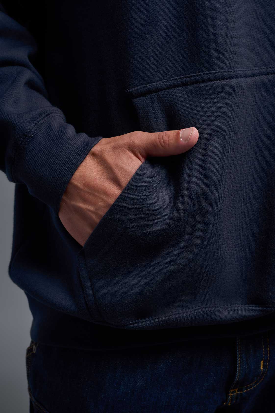 A male model wearing the unisex Americana 3-End Fleece Pullover Hoodie in the color navy, made from 75% combed ring-spun USA-grown cotton and 25% polyester, showing the relaxed fit from a close-up pocket view. This premium fleece hoodie is Made in the USA. #navy