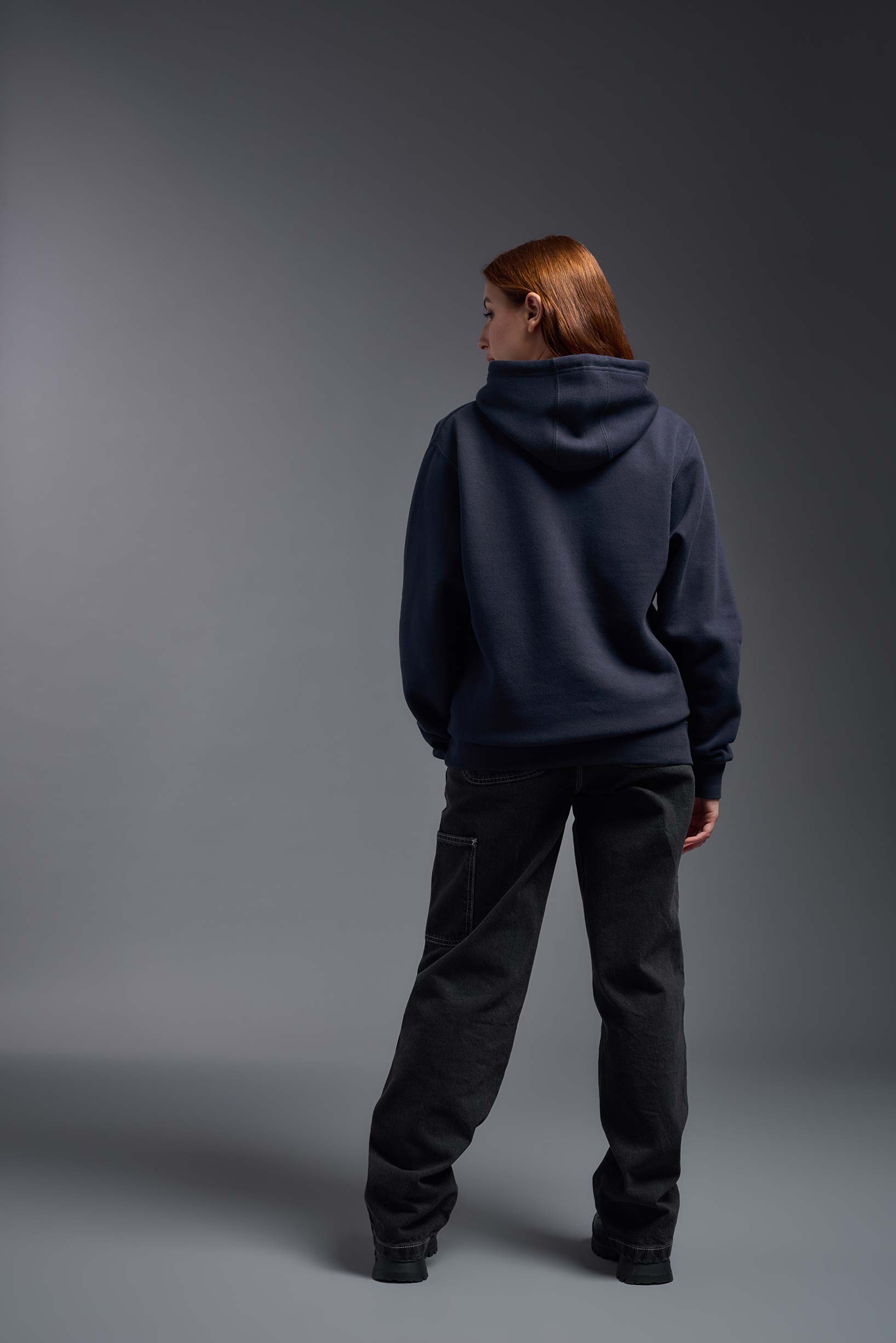 A female model wearing the unisex Americana 3-End Fleece Pullover Hoodie in the color navy, made from 75% combed ring-spun USA-grown cotton and 25% polyester, showing the relaxed fit from a back view. This premium fleece hoodie is Made in the USA. #navy
