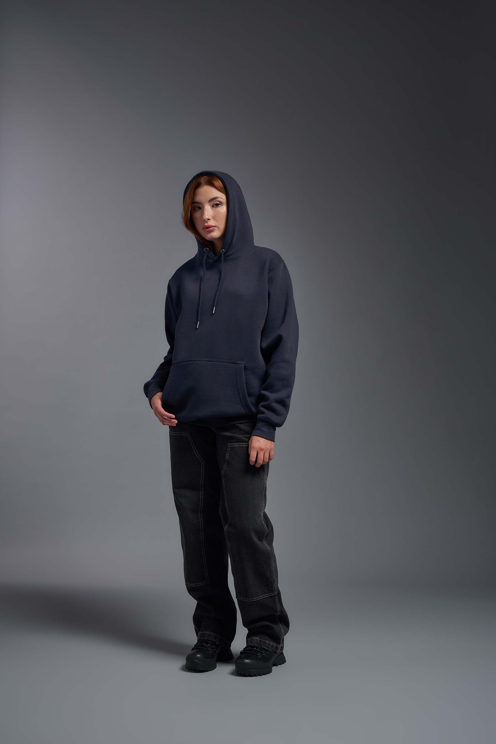 A female model wearing the unisex Americana 3-End Fleece Pullover Hoodie in the color navy, made from 75% combed ring-spun USA-grown cotton and 25% polyester, showing the relaxed fit from a front view with hood up. This premium fleece hoodie is Made in the USA. #navy