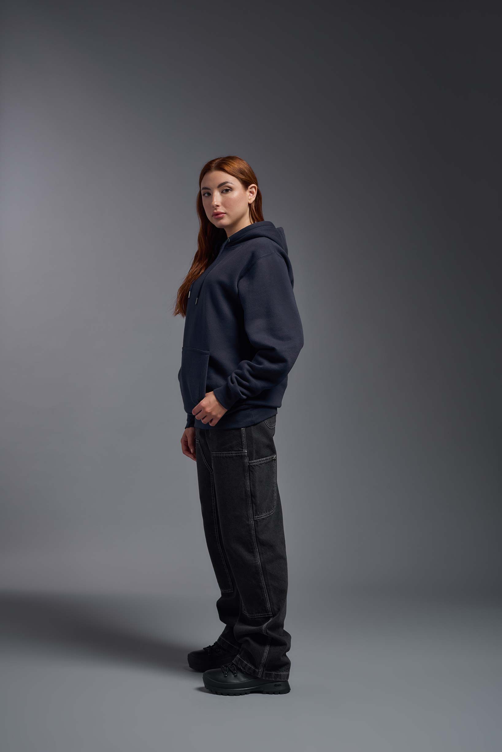 A female model wearing the unisex Americana 3-End Fleece Pullover Hoodie in the color navy, made from 75% combed ring-spun USA-grown cotton and 25% polyester, showing the relaxed fit from a side view. This premium fleece hoodie is Made in the USA. #navy