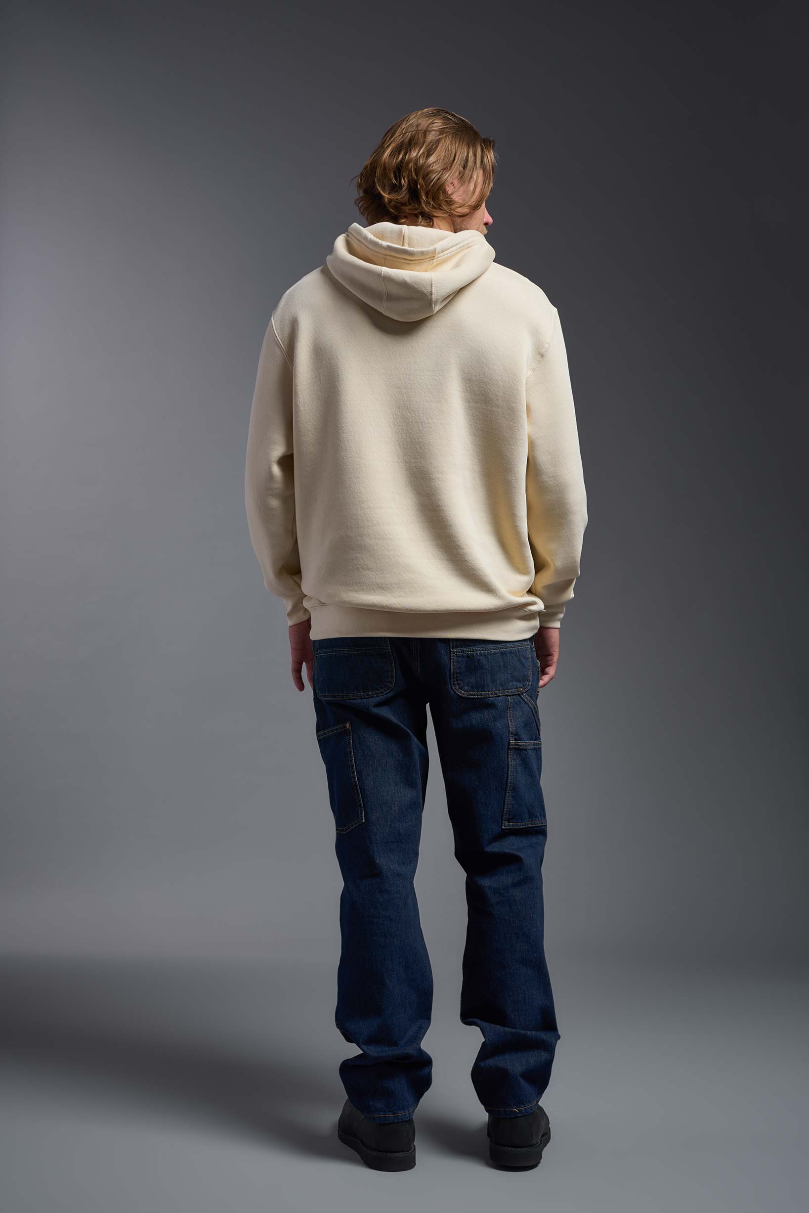 A male model wearing the unisex Americana 3-End Fleece Pullover Hoodie in the color sand, made from 75% combed ring-spun USA-grown cotton and 25% polyester, showing the relaxed fit from a back view. This premium fleece hoodie is Made in the USA. #sand