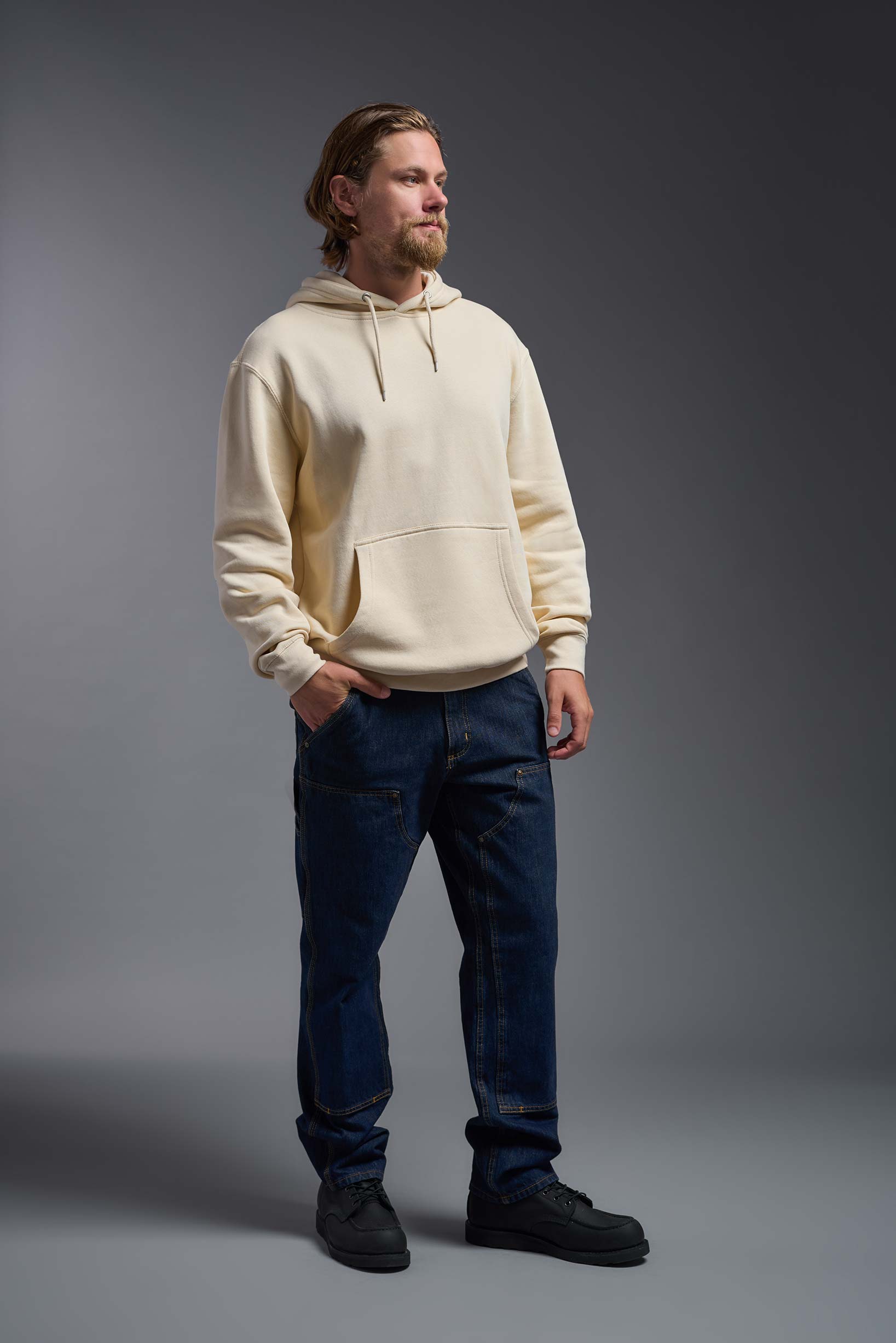 A male model wearing the unisex Americana 3-End Fleece Pullover Hoodie in the color sand, made from 75% combed ring-spun USA-grown cotton and 25% polyester, showing the relaxed fit from a side view. This premium fleece hoodie is Made in the USA. #sand