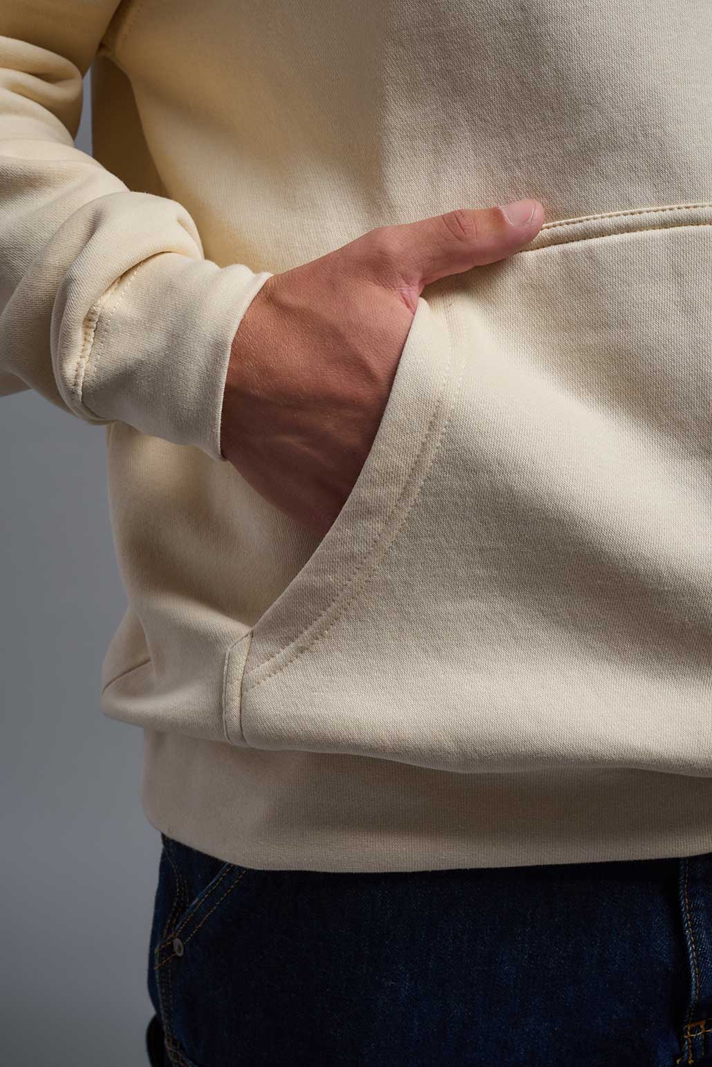 A male model wearing the unisex Americana 3-End Fleece Pullover Hoodie in the color sand, made from 75% combed ring-spun USA-grown cotton and 25% polyester, showing the relaxed fit from a close-up pocket view. This premium fleece hoodie is Made in the USA. #sand