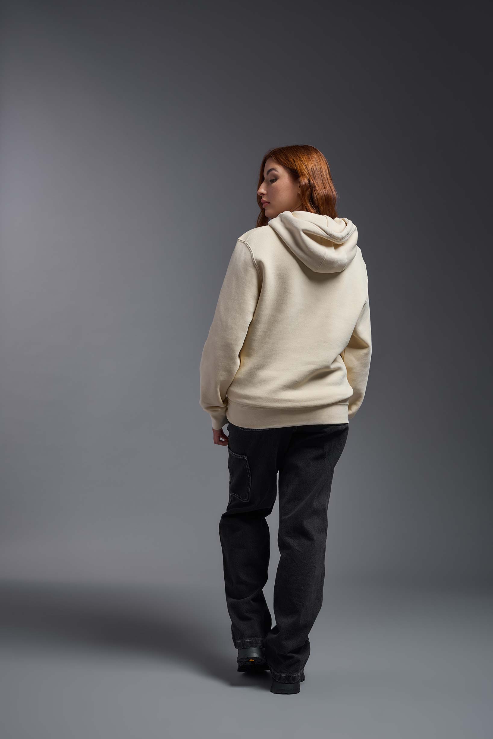 A female model wearing the unisex Americana 3-End Fleece Pullover Hoodie in the color sand, made from 75% combed ring-spun USA-grown cotton and 25% polyester, showing the relaxed fit from a back view. This premium fleece hoodie is Made in the USA. #sand