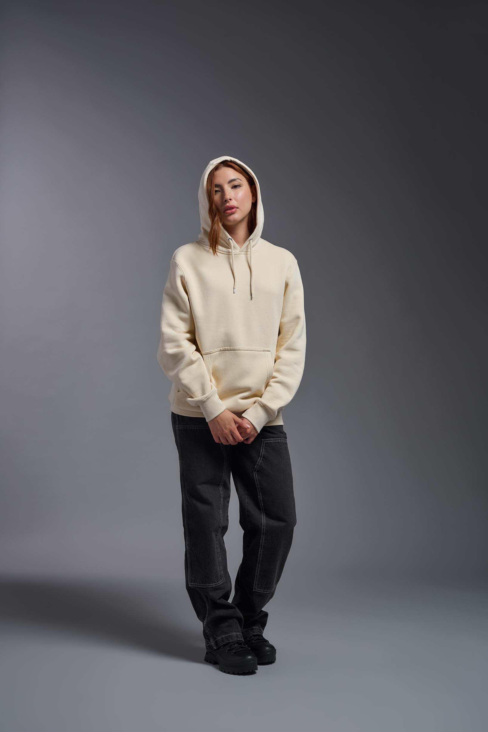 A female model wearing the unisex Americana 3-End Fleece Pullover Hoodie in the color sand, made from 75% combed ring-spun USA-grown cotton and 25% polyester, showing the relaxed fit from a front view with hood up. This premium fleece hoodie is Made in the USA. #sand