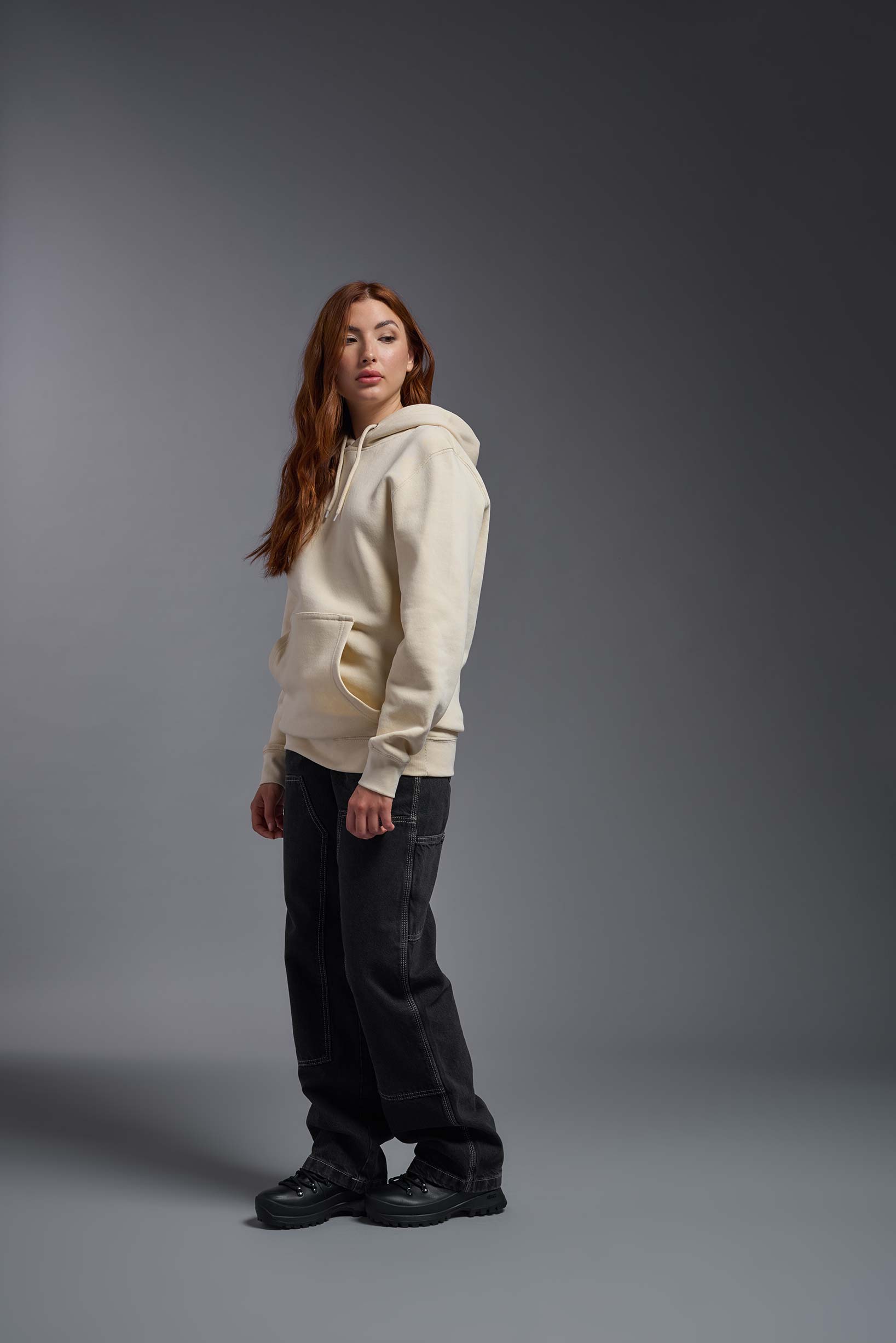 A female model wearing the unisex Americana 3-End Fleece Pullover Hoodie in the color sand, made from 75% combed ring-spun USA-grown cotton and 25% polyester, showing the relaxed fit from a side view. This premium fleece hoodie is Made in the USA. #sand