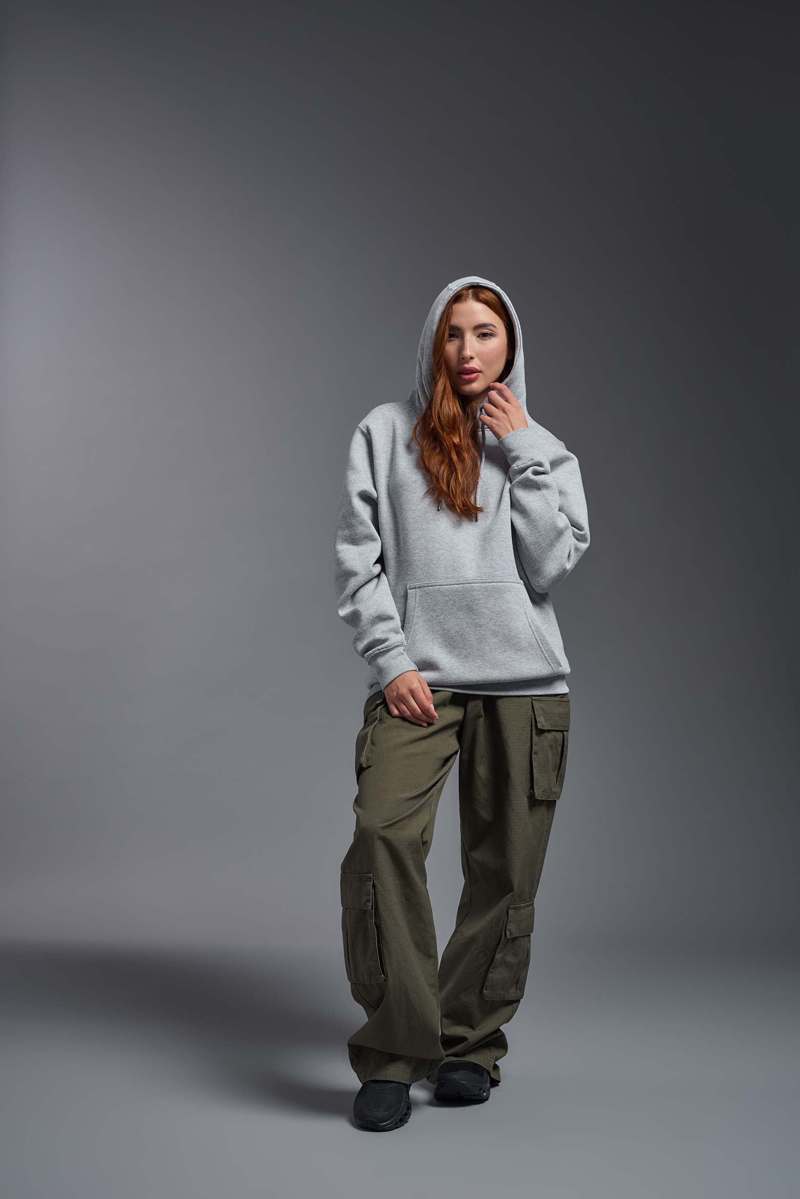 A female model wearing the unisex Americana 3-End Fleece Pullover Hoodie in the color sport gray, made from 75% combed ring-spun USA-grown cotton and 25% polyester, showing the relaxed fit from a front view with hood up. This premium fleece hoodie is Made in the USA. #sport-gray