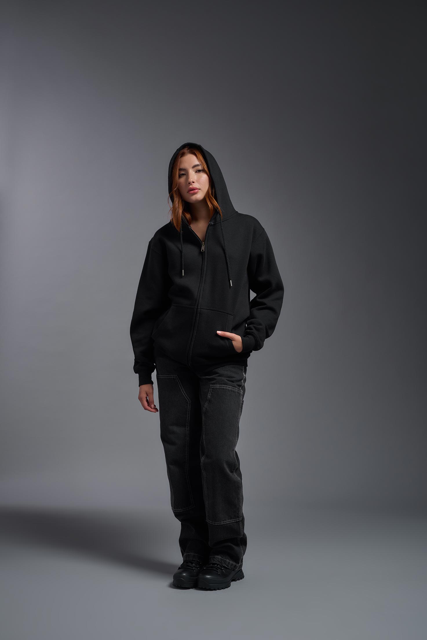 A female model wearing the unisex Americana 3-End Fleece Full Zip Hoodie in the color black, made from 75% combed ring-spun USA-grown cotton and 25% polyester, showing the relaxed fit from a front view with her hood up. This premium fleece hoodie is Made in the USA. #black