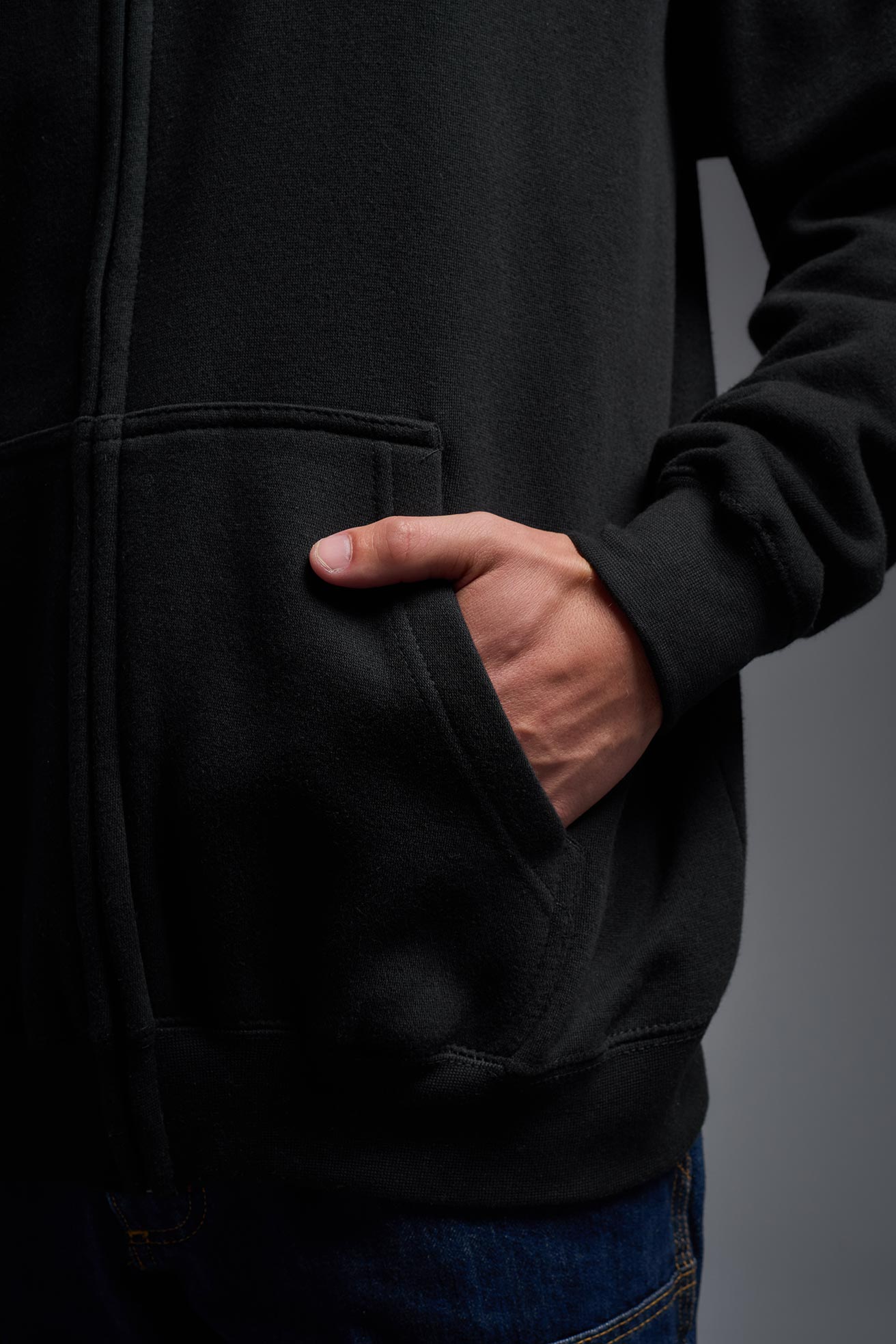 A male model wearing the unisex Americana 3-End Fleece Full Zip Hoodie in the color black, made from 75% combed ring-spun USA-grown cotton and 25% polyester, showing the relaxed fit from a pocket view. This premium fleece hoodie is Made in the USA. #black