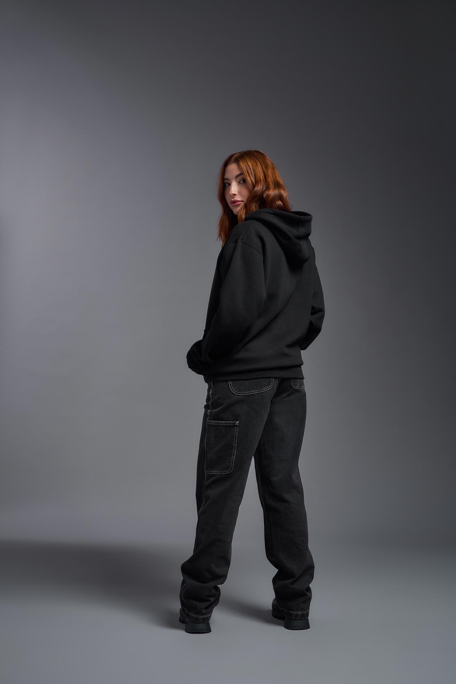 A female model wearing the unisex Americana 3-End Fleece Full Zip Hoodie in the color black, made from 75% combed ring-spun USA-grown cotton and 25% polyester, showing the relaxed fit from a back view. This premium fleece hoodie is Made in the USA. #black