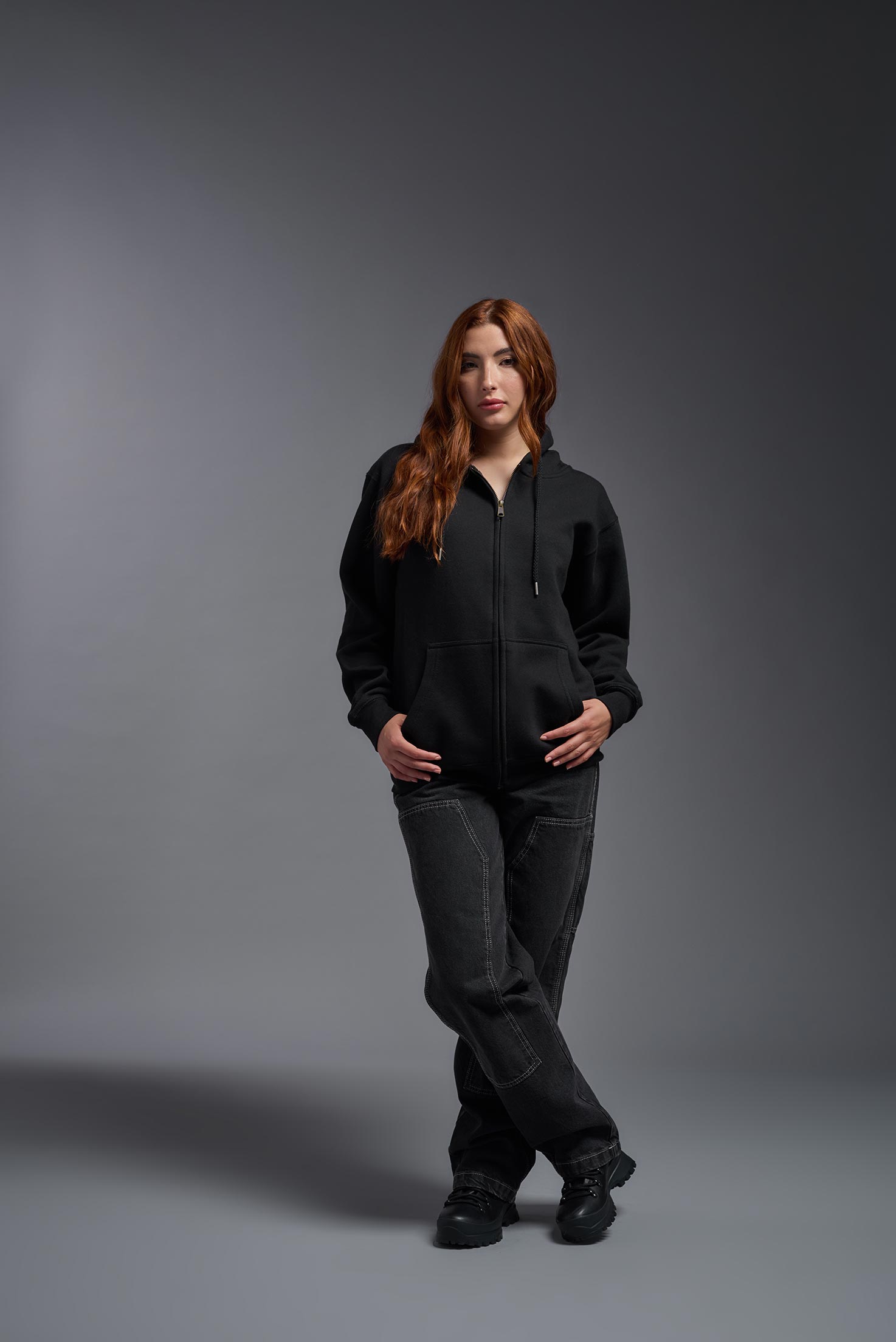 A female model wearing the unisex Americana 3-End Fleece Full Zip Hoodie in the color black, made from 75% combed ring-spun USA-grown cotton and 25% polyester, showing the relaxed fit from a front view. This premium fleece hoodie is Made in the USA. #black