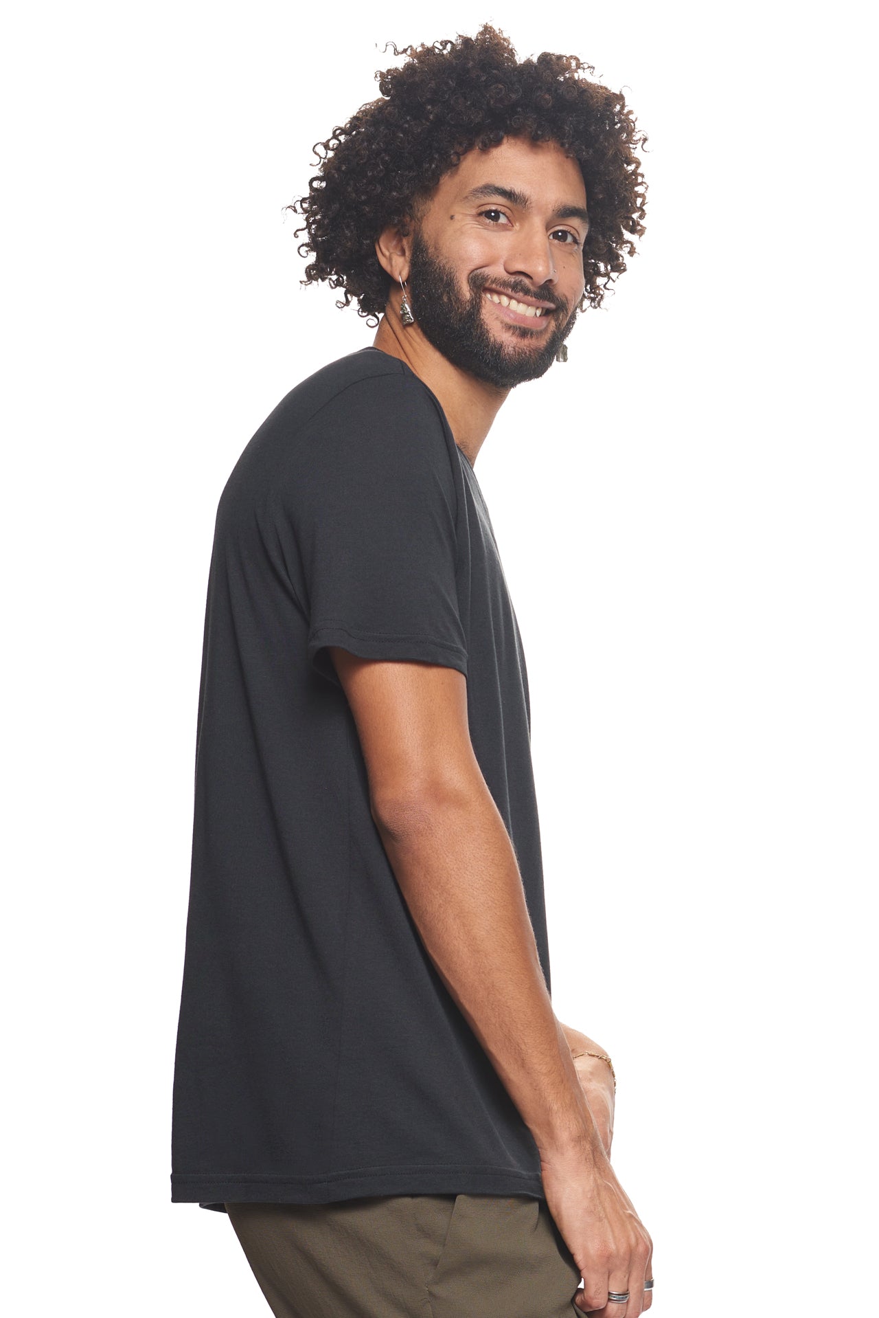 A male model with curly hair wearing the Men's MoCA V-Neck Tee in the color black, made from a luxurious blend of 50% LENZING Modal and 50% Combed Ring-Spun Cotton, showing the relaxed fit from a side view. This fashion-forward, more sustainable t-shirt is elevated, eco-friendly, made in the USA, and available for bulk purchase. #black